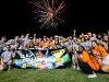  large group of Wichita State students in gray and orange shirts pose together at night holding a “Clash of the Colleges Champions” banner. Fireworks light up the sky above them as they smile and celebrate their win.