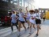 A group of energetic students, some wearing College of Health Professions shirts, run and laugh together during a campus event. They hold phones and wear sunglasses and beads, with the sun shining behind them.