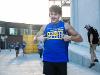 A student in a blue “College of Applied Studies” tank top points proudly to his shirt while smiling. Other students in similar shirts are visible running in the background during a campus event.