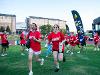 Students wearing red “Barton School of Business” shirts run across a grassy field with big smiles during Clash of the Colleges. Behind them, more students in matching shirts and red face paint follow, with colorful event flags in the background.