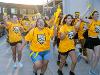 A large group of Wichita State students in bright yellow shirts with WuShock logos cheer and run forward during a campus event. Many are holding cowbells, noisemakers, or vuvuzelas and wearing face paint and bandanas.