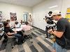 Students gather in a classroom for a group project discussion while a film crew records the session.