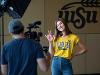 A Wichita State student smiles and waves during an on-camera interview, with the WSU logo in the background.