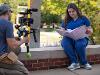 A student in scrubs studies from a pink binder labeled “Intro to Micro” while being filmed outdoors.