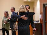 Bobby Gandu smiles while hugging a woman during a memorial service reception. He holds a glass of wine, and other guests are visible in the background.
