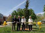 Four people stand in front of a tall evergreen tree, cutting a yellow ribbon during a dedication ceremony. They are holding large ceremonial scissors under a clear blue sky.