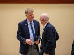 Rick Case, wearing a blue suit and tie, speaks with another attendee at a memorial service. They are standing in conversation in a formal indoor setting.