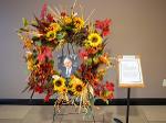 A memorial wreath made of autumn flowers and leaves surrounds a photo of Dr. Jim Rhatigan. A printed tribute stands on an easel beside the wreath.
