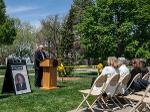 A man stands at a podium outdoors, speaking to a seated audience during a tree dedication ceremony. A large photo of Dr. Jim Rhatigan is displayed beside him, and yellow ribbons mark the newly planted tree.