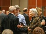Beverly Rhatigan, wearing a patterned jacket, warmly greets a man in a suit during a memorial service. People gather and talk in the background.
