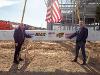 Wichita State's John Tomblin and President Rick Muma stand on either side of a signed steel beam at the construction site of the Hub for Advanced Manufacturing and Research, with the building framework and an American flag in the background.