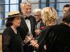 A group of people in formal attire stand and talk while holding wine glasses at a social event. Large windows in the background reveal an urban view.