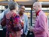 Three people stand outdoors at the Ulrich50 Gala, holding wine glasses and talking. One wears a pink jacket, another a blue jacket and the third a colorful floral-patterned outfit. A railing and some structures are visible in the background.
