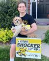 A student poses outside of their home beside a Future Shocker yard sign while holding their dog.
