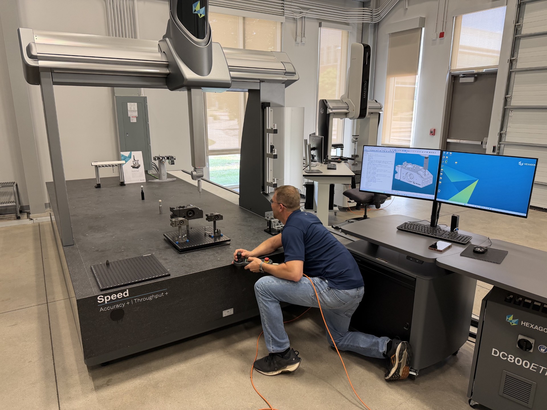 The image shows a person working in a modern industrial metrology lab. The individual is kneeling beside a large coordinate measuring machine (CMM) with the brand “Hexagon” visible on the equipment. The machine has a wide granite base labeled “Speed – Accuracy + Throughput,” and several metal components and fixtures are arranged on its surface for measurement.
The person is holding a controller connected by an orange cable, appearing to adjust or operate the CMM. Nearby is a workstation with dual monitors displaying CAD models and measurement software. The workspace includes a keyboard, mouse, and additional computer equipment on a sleek, dark workstation cabinet labeled “DC800ET.”
The room itself has high ceilings, exposed conduit, large windows, and polished concrete floors, giving it a clean, industrial, and high-tech environment. Multiple pieces of precision equipment and workstations are positioned around the room, indicating a professional engineering or manufacturing measurement facility.