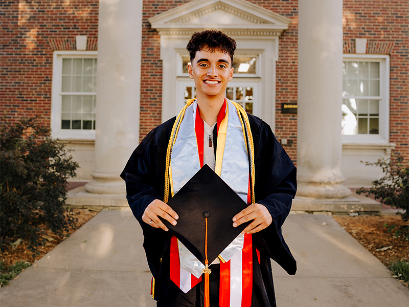 Photo of Michael Boydo standing in front of a Wichita State building  wearing their graduation cap and gown, graduation stole, and honors cord. 