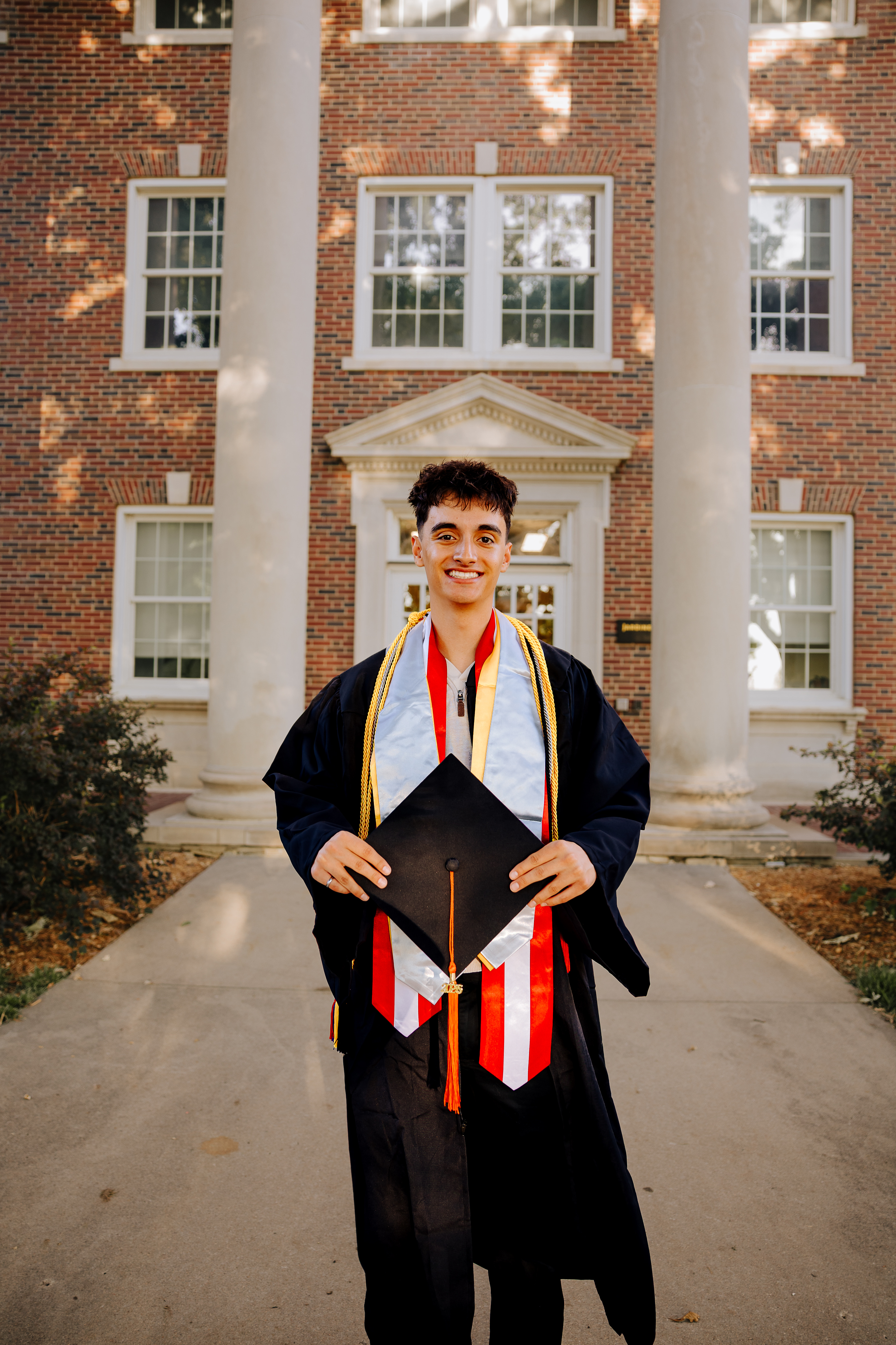 Photo of Michael Boydo standing in front of a Wichita State building  wearing their graduation cap and gown, graduation stole, and honors cord. 