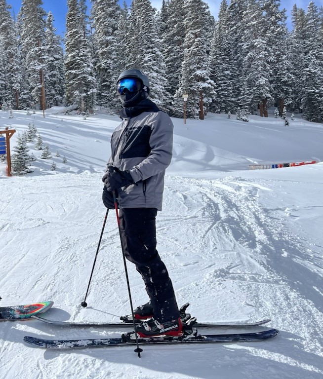Photo of Justen Lee on a ski slope, in ski gear, and in front of snow covered trees.