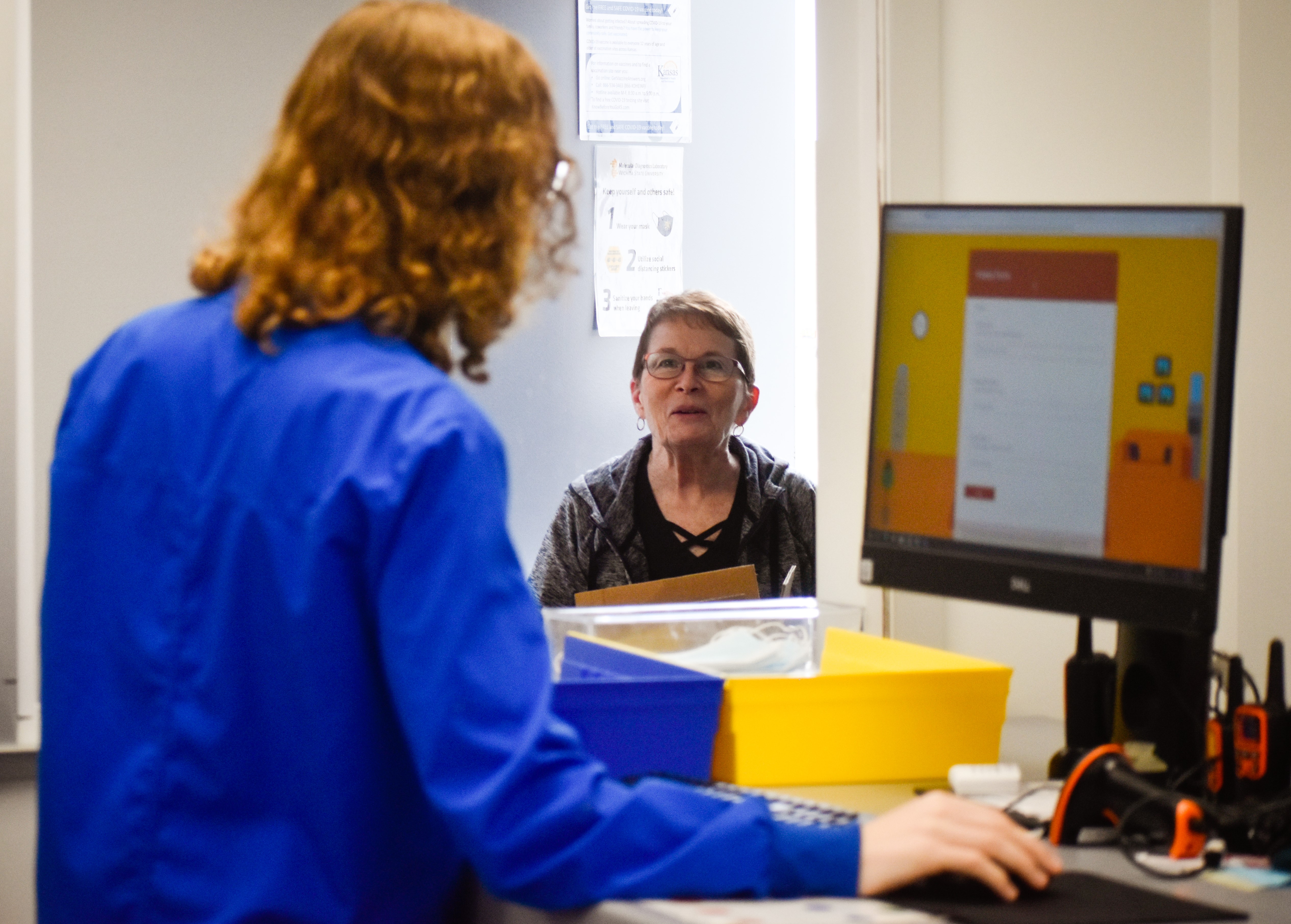 An woman goes to drop off a sample at MDL's intake drop off window. Another woman in a blue lab coat is logging the drop off on her computer.