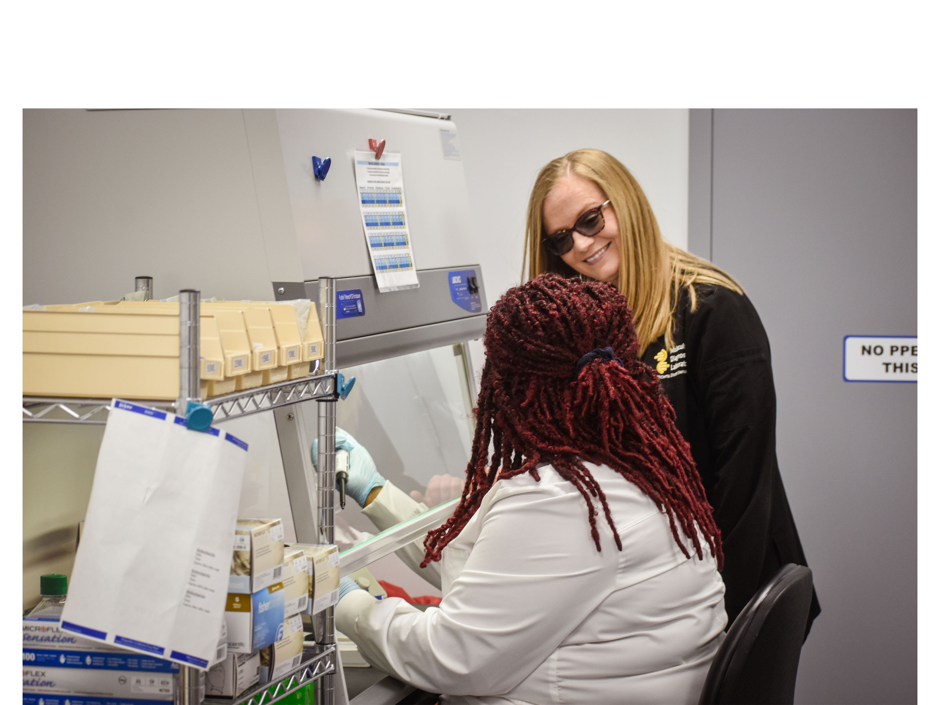 A woman in PPE sits under a medical hood to pipetting a sample while another woman stands facing her smiling.