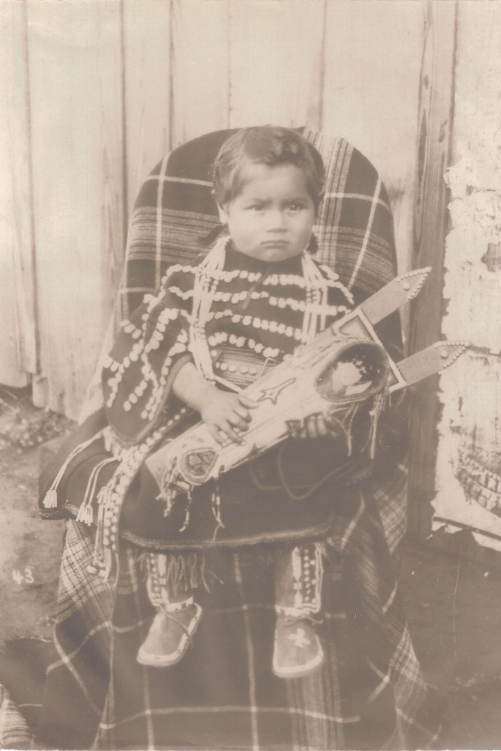 Small girl wearing a decorated dress while holding porcelain faced doll.