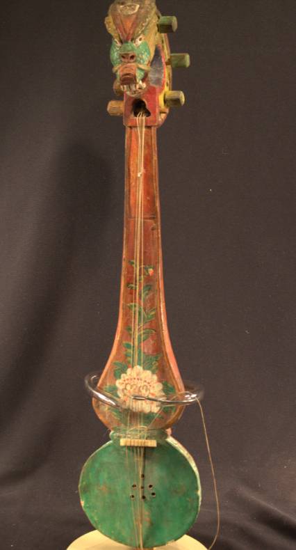 A string instrument that has a base shaped like an eight. The head of the instrument is carved to look like the head of a dragon. The base is painted green with four gold swastikas. A flower starts at the base of the instrument and makes its way up the neck.