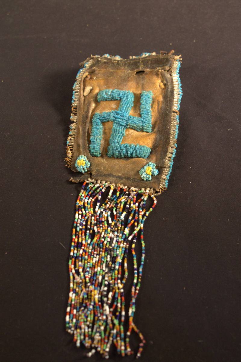 A small brown leather drawstring bag that is starting to deteriorate. Multicolored beads hang from the bottom, blue beads create a border around the edge, two flowers made from blue and yellow beads are located in the bottom corners, and in the center is a swastika made of blue beads.