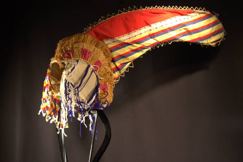 Headdress is red conical part with blue, red, and yellow stripes with decorative flower patterns.
