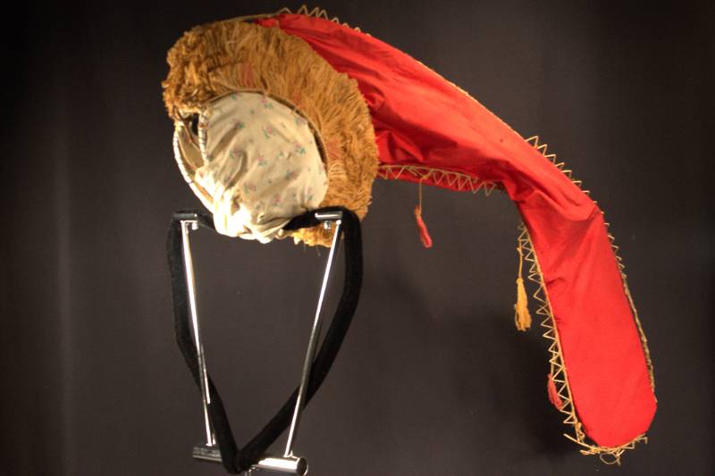 Headdress is red conical shaped fabric with wooden triangle decoration along perimer.  Fabric tassels hanging from conical part headdress part has fibers around outside the fabric covering the hat part is white with pink roses.