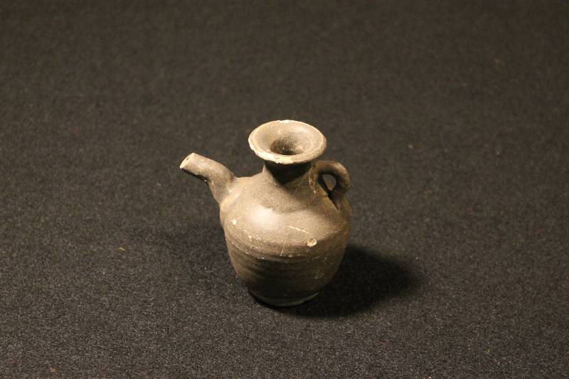 Medium Sawankhalok ware spouted jarlet. Over-all brown glaze covers pot. Extended neck is fitted with an ear-shaped loop handle and the body with a pouring spout. Opening center top, finished with flared lip.