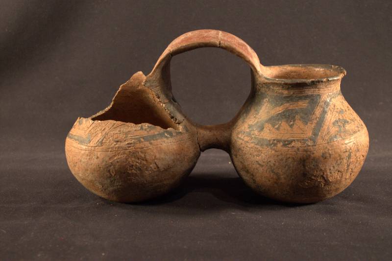 Double-jar vessel is a red/brown color with a black geometric design. Two jars are connected by a handle in the center and the left jar is heavily damaged with the top missing.