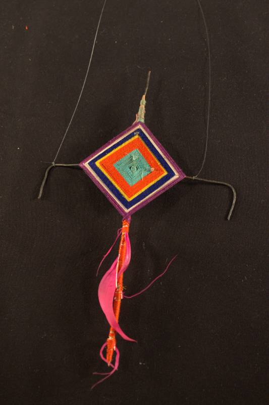 Ornamental head piece made from metal wire and wrapped with colorful string (light blue, red, yellow, blue, white, and purple) the top has red string wrapped around it with pink feathers.