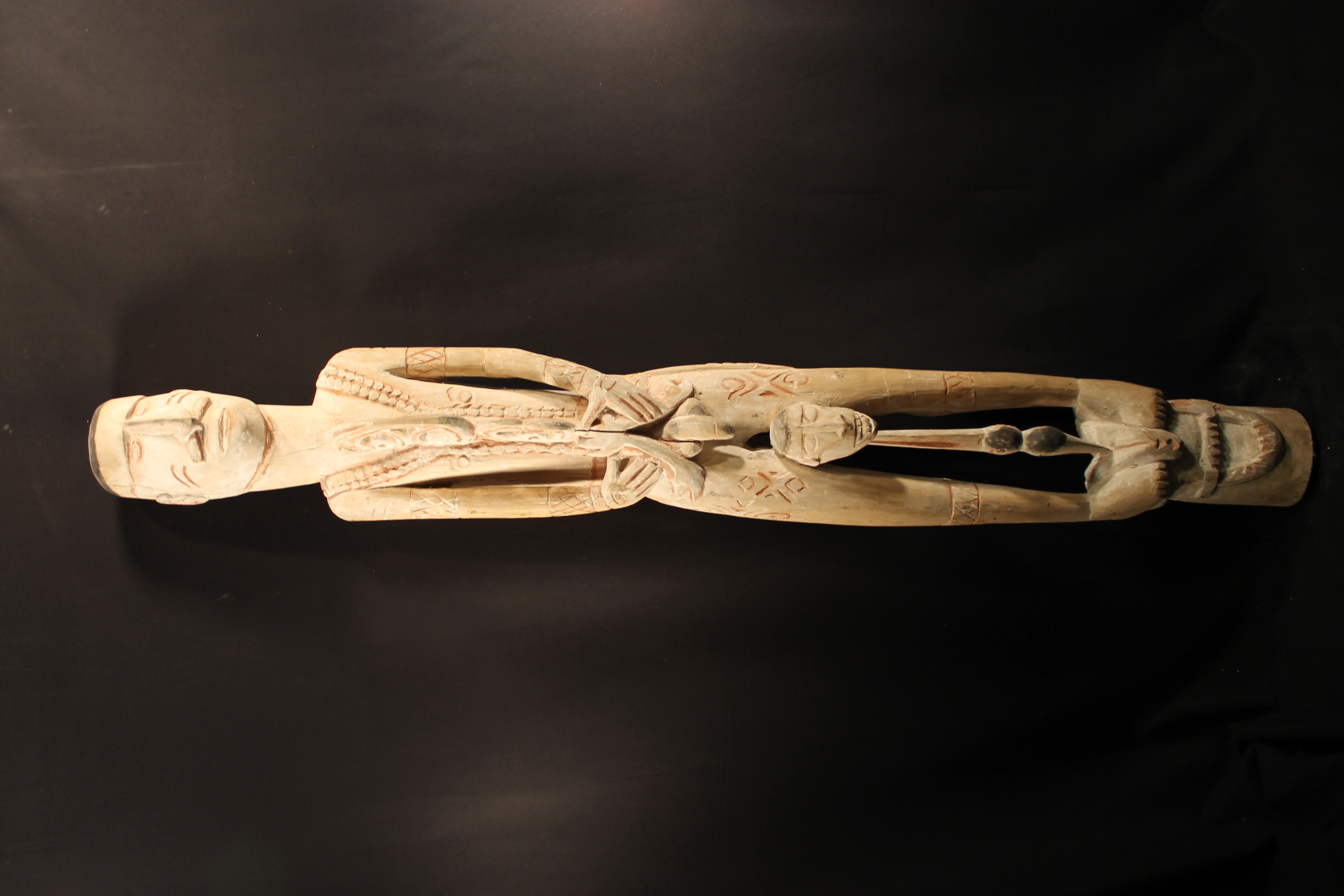 Male ancestor figure wearing vest and armbands is standing on a carving of a mouth. Small head carved sitting on top of a carved bird’s head. 