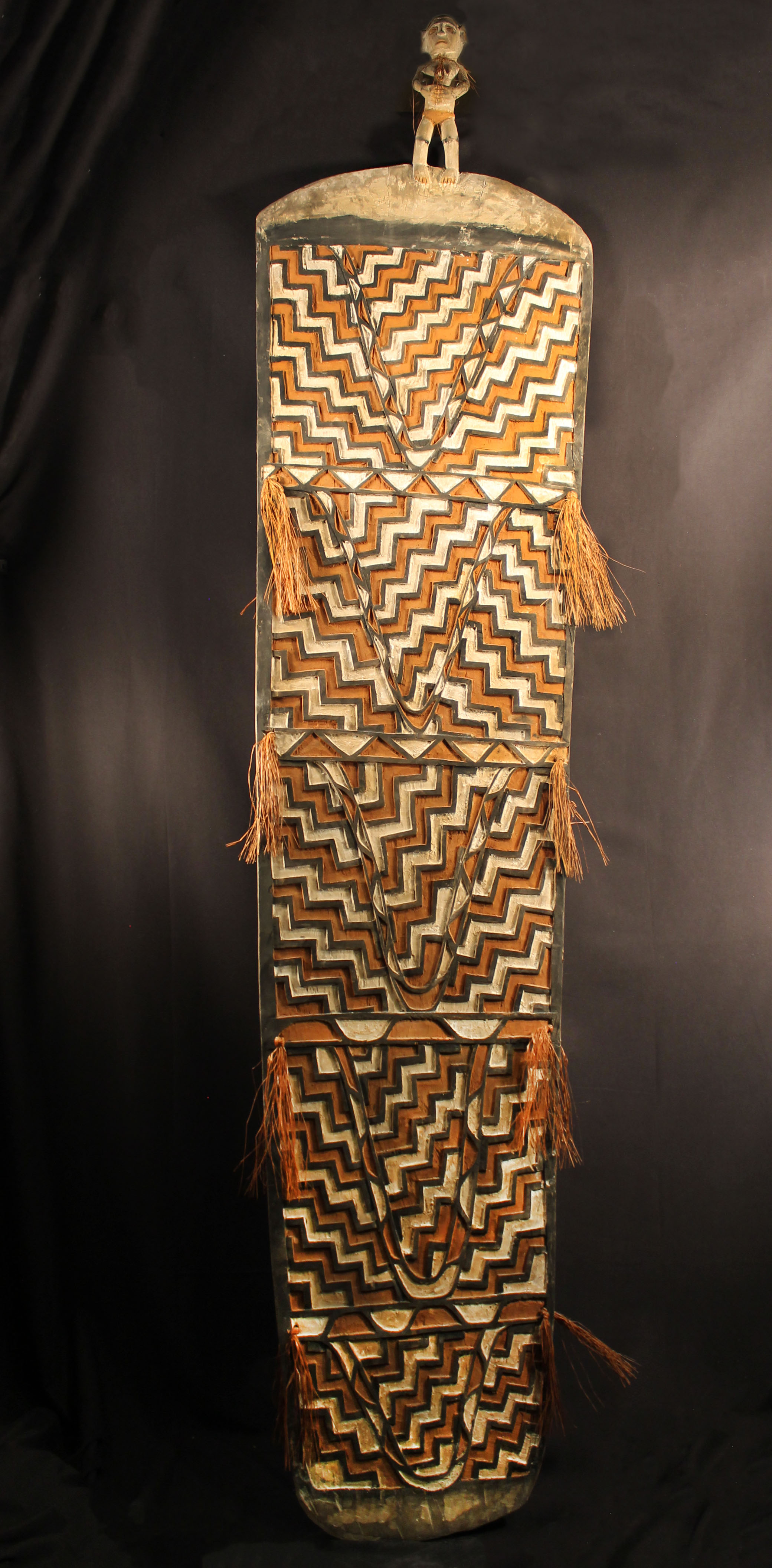Wood carved large shield with intricate geometric designs with tassels on the side. Human figure sits on the top. Red, white, and black pigment used to decorate carvings