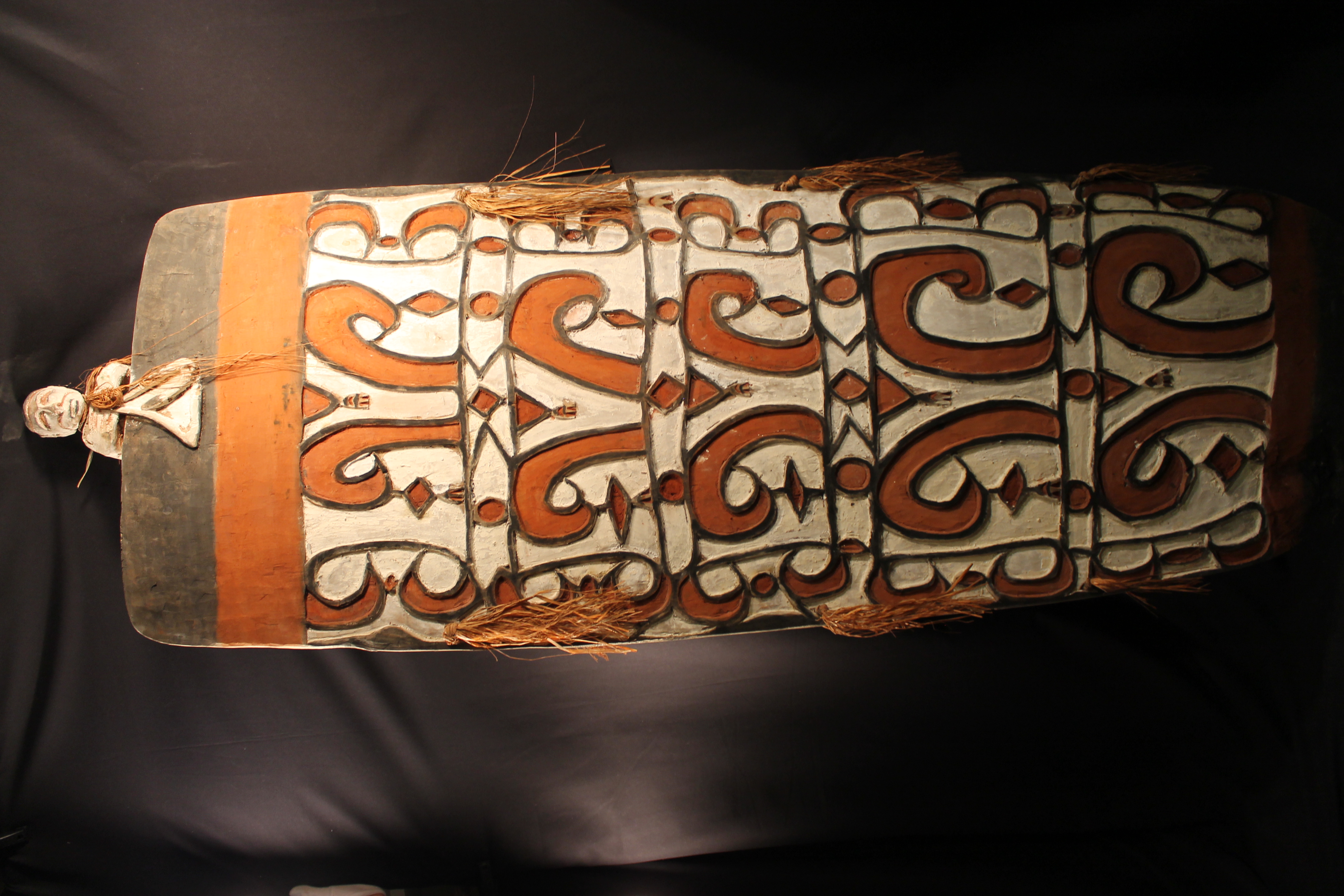 Carved wooden shield with an ancestral figure at the top with two tassels around the neck and on the sides. The carved design is decorated with black, white, and red pigment.   