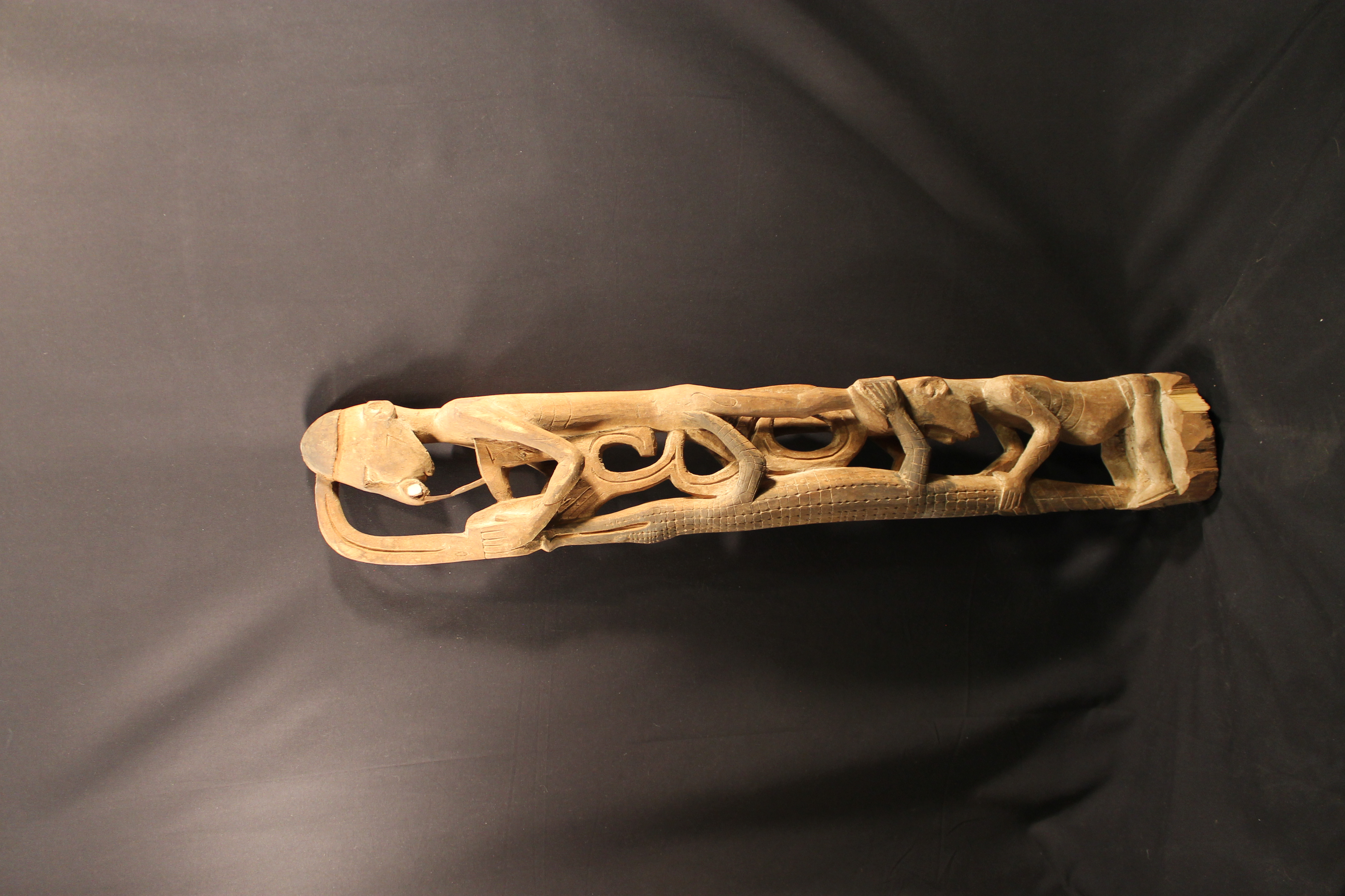 Wooden carving of a seated woman and male, who is wearing a nose piece and is standing on her head. They are both holding a large crocodile