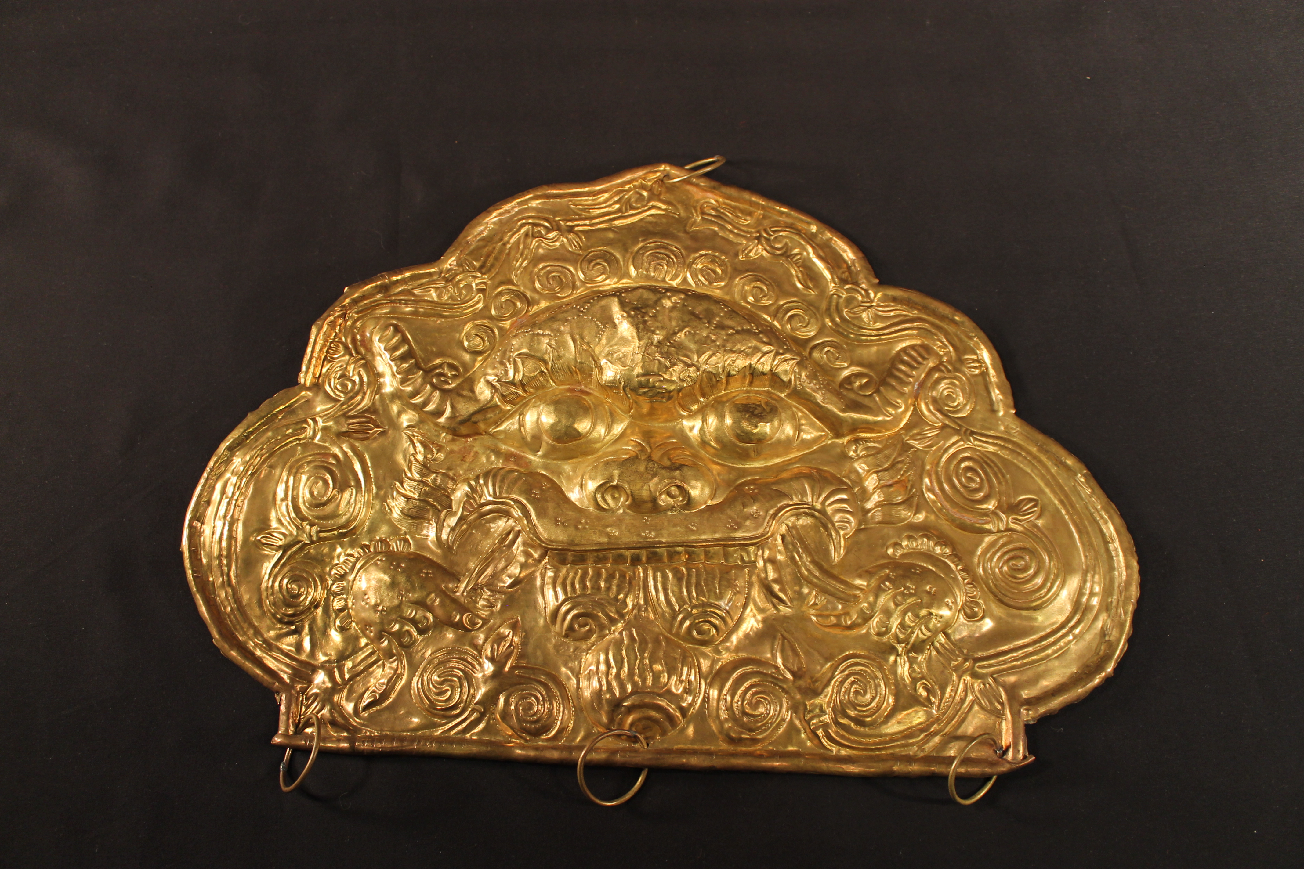 The plaque depicts the Kirtimukha monster, with large eyes, fangs, horns, and human-like hands. Swirling designs surround the creature.