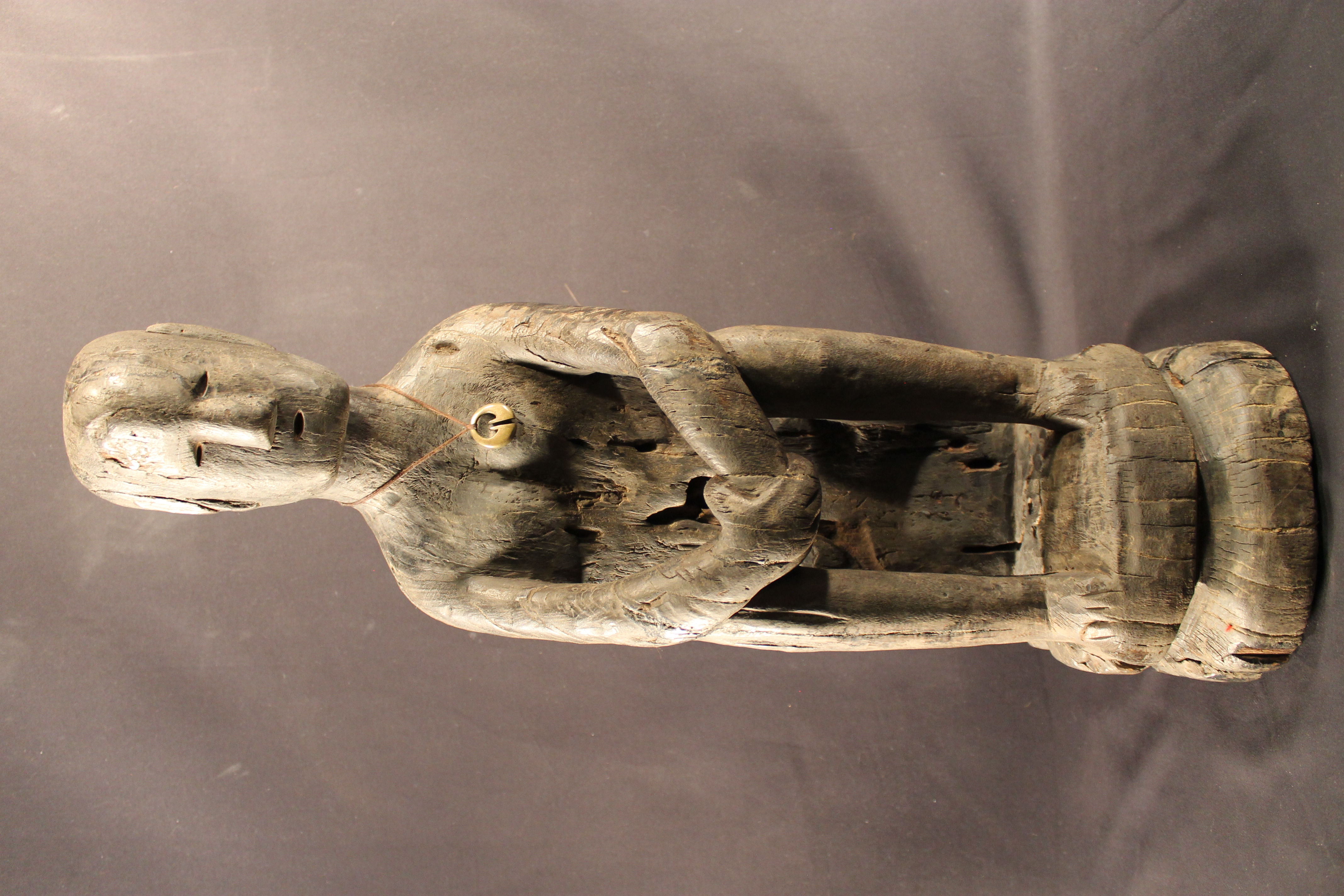 Wood-carved statue of a man sitting. The man is holding his hand and is wearing a necklace. The statue has various cracks on the wood and small insect holes in the wood.