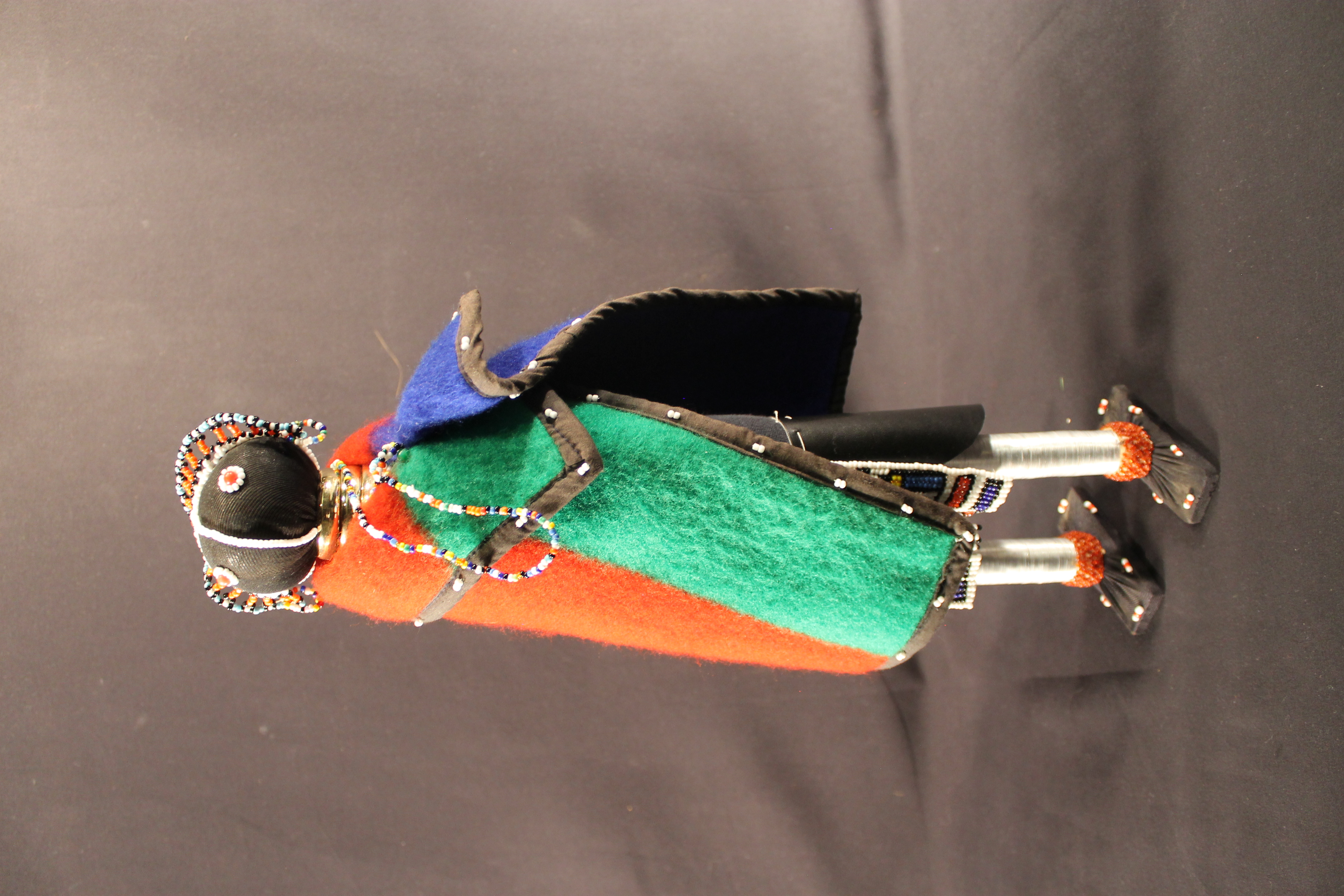 The doll has beaded features and headdress, gold neck rings, multicolored beaded necklace and earrings. There is white beading on the red, green, and blue cap. It also has a black sheath with a multicolored patterned beaded apron.