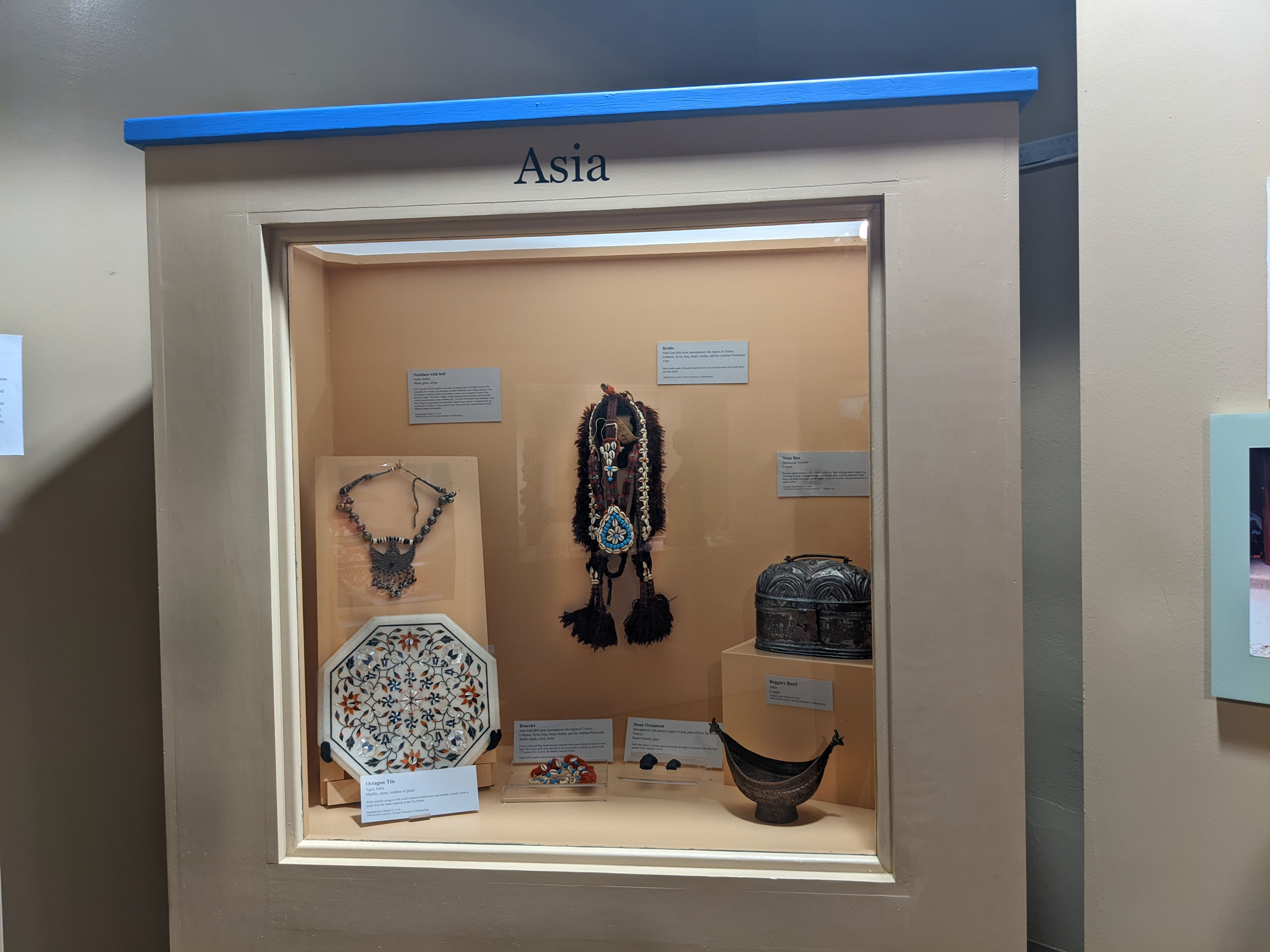 Case with orange background. Objects include octagon tile, necklace, bracelet, ornaments, bridle, and metal box. Labels attached to the back wall and on the bottom surface.