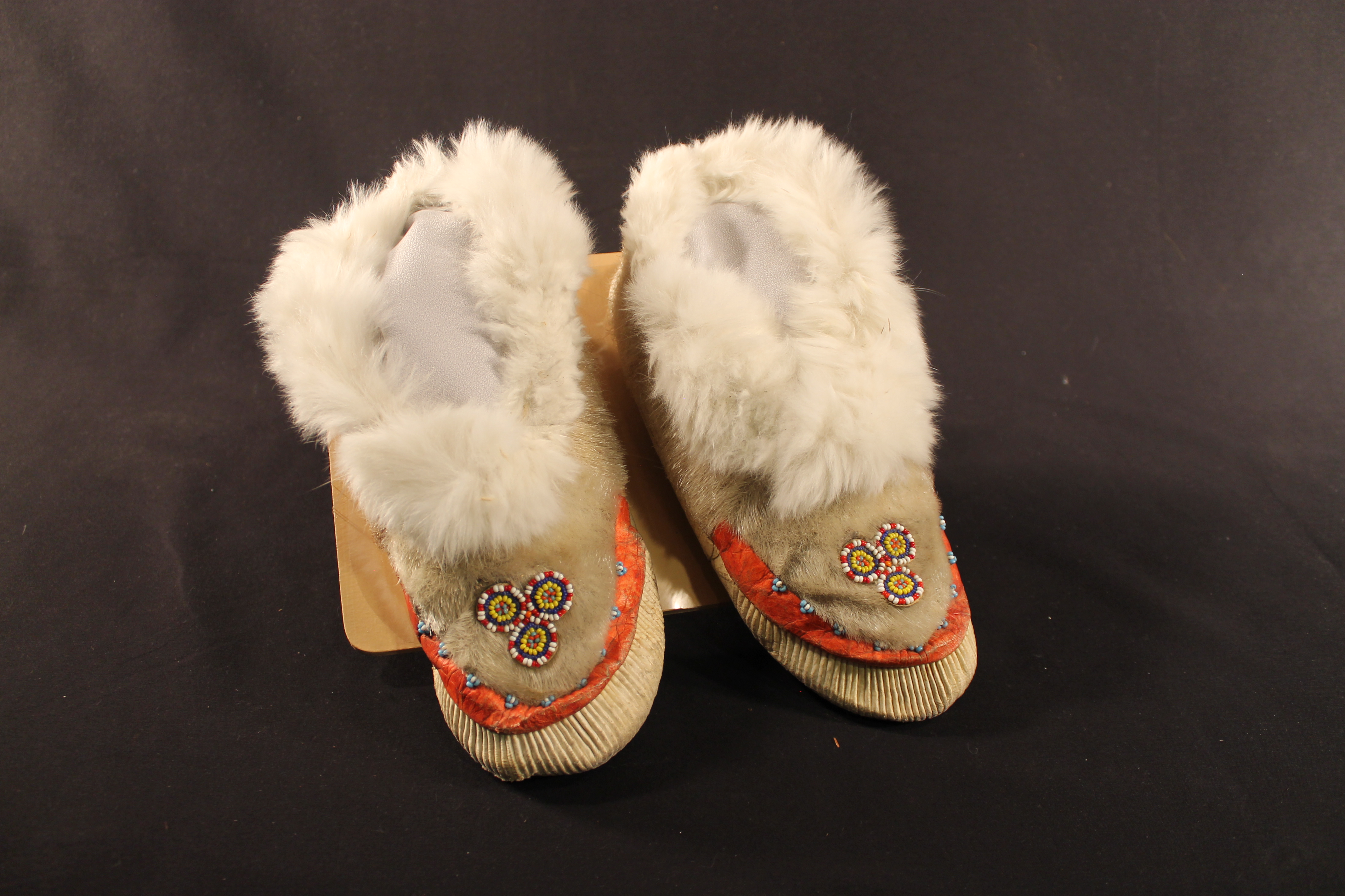 The pair of moccasins are made of furry hide on the interior and exterior. The tie and sole of the shoes is made up of white pleated leather. On top of that is a band of red leather decorated with blue and white beads. On the center of the shoes is a bead design of three circles touching each other.