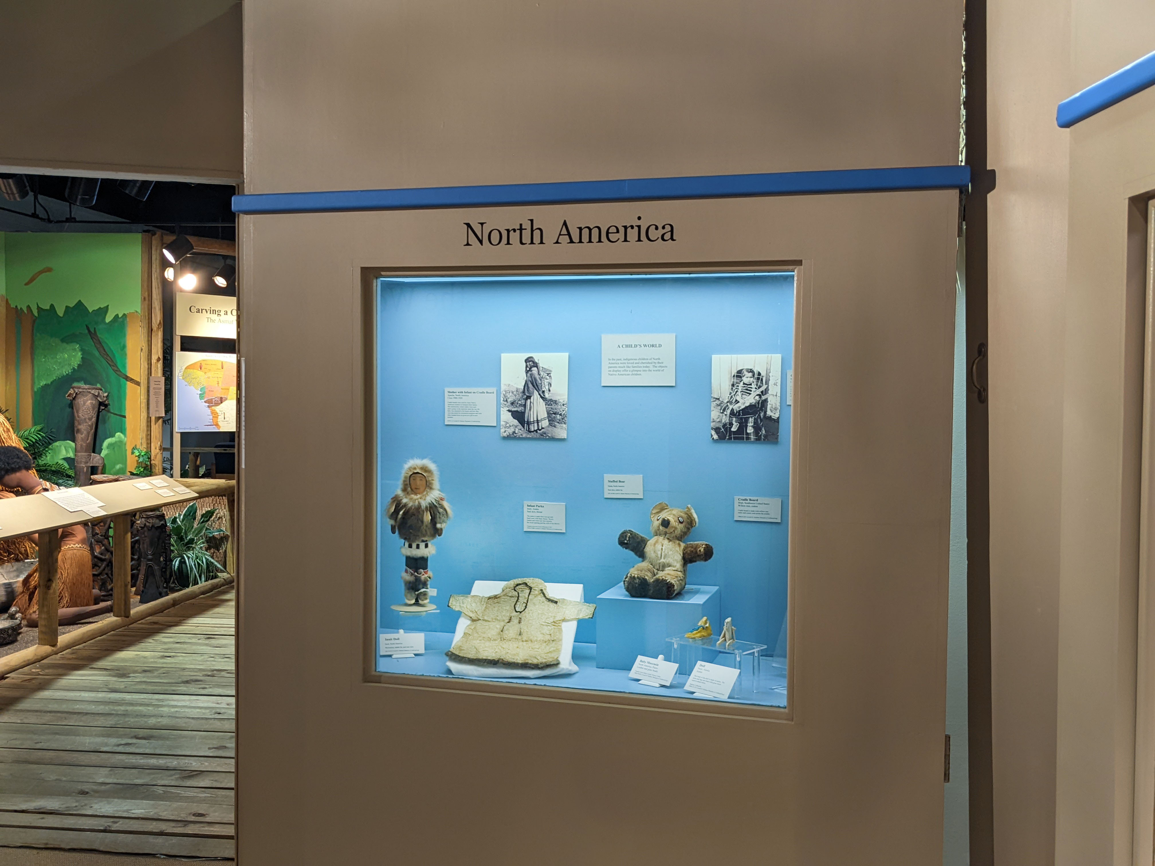 The case has a blue background with items displayed below including a doll, small jacket, stuffed bear, small moccasin, small figure, and cradleboard. Label information is displayed on the wall behind the items, as well as in front of them. The case also has two photographs. The first is of a mother wearing a cradleboard that is supporting her infant on her back. The other is of a child attached to a cradleboard sitting down and holding a doll.