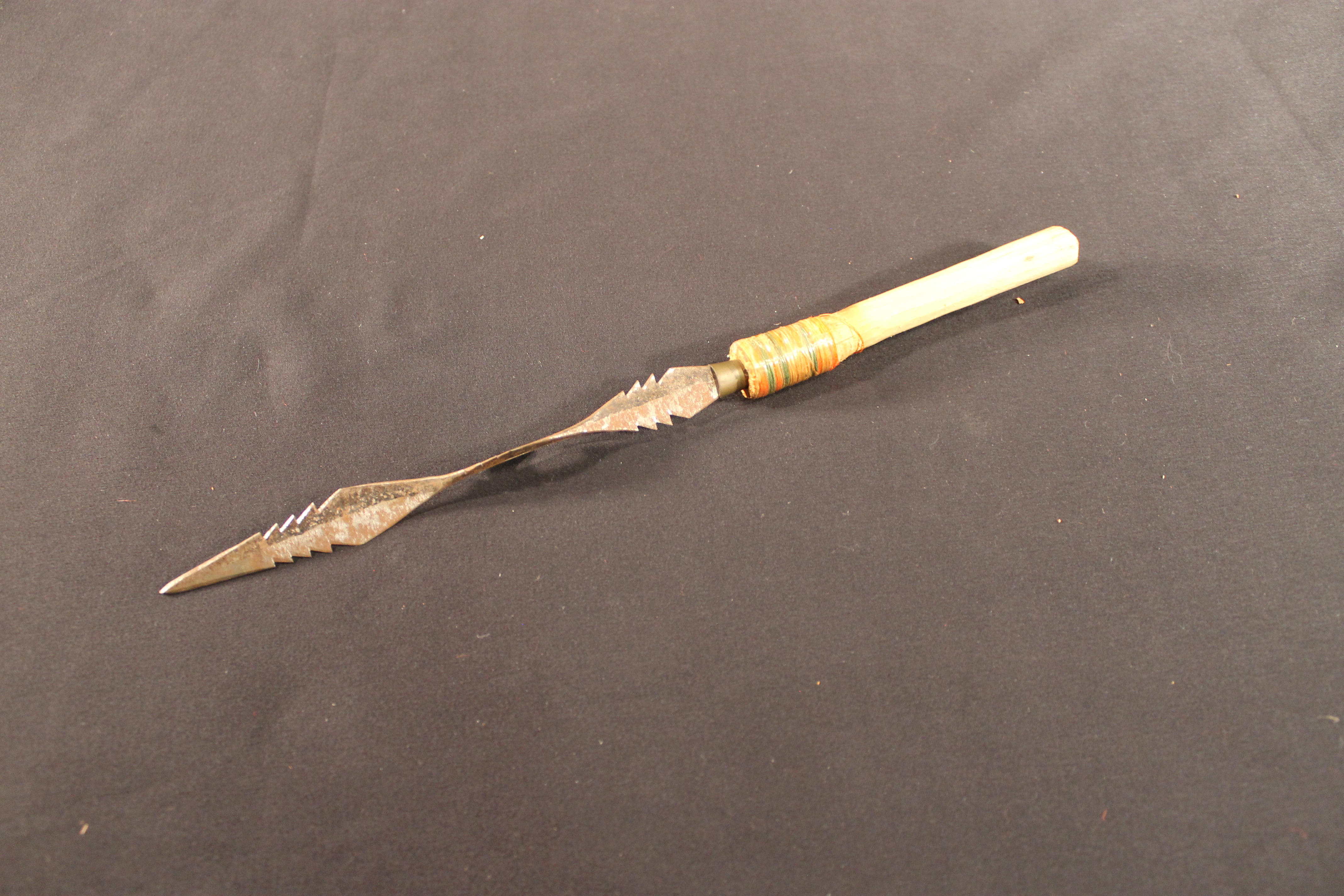 Dart made of metal and wood wrapped with bands of orange, green, yellow, and off-white threads.