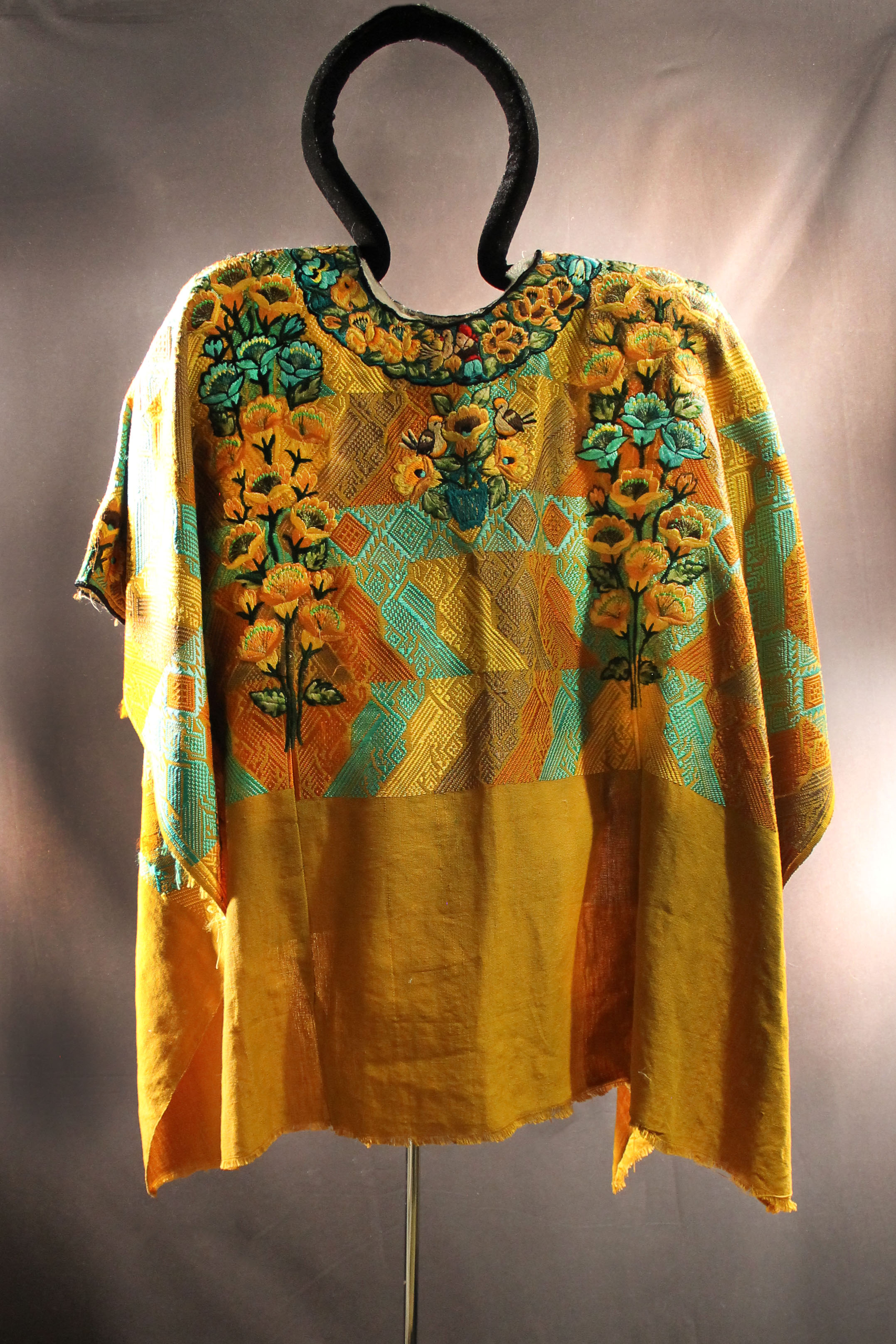 Huipil has an orange, blue, brown, and yellow woven pattern.  Flowers as the main design with birds in the middle.