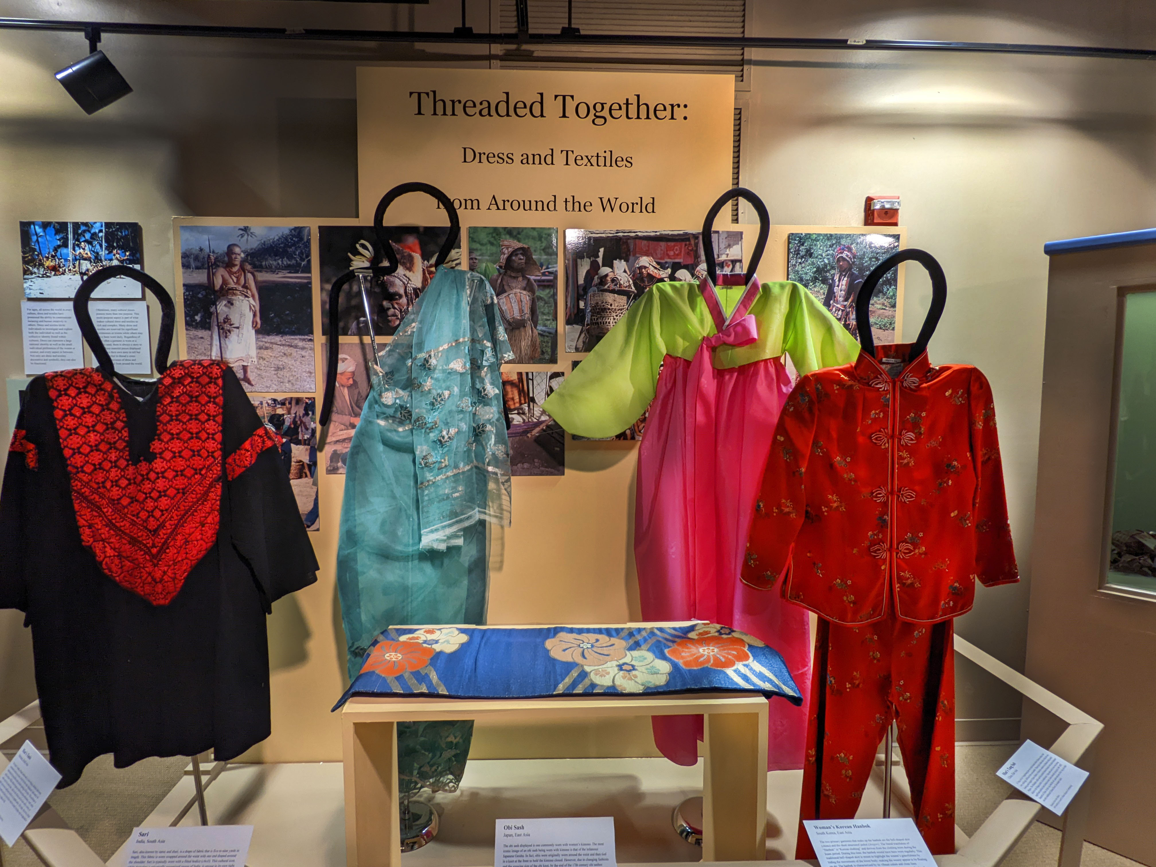 Open case with yellow background with mannequins and pedestals with garments on them. Behind them is a display of images of people wearing traditional costumes. The labels are attached to a rail that sits on the front of the display.