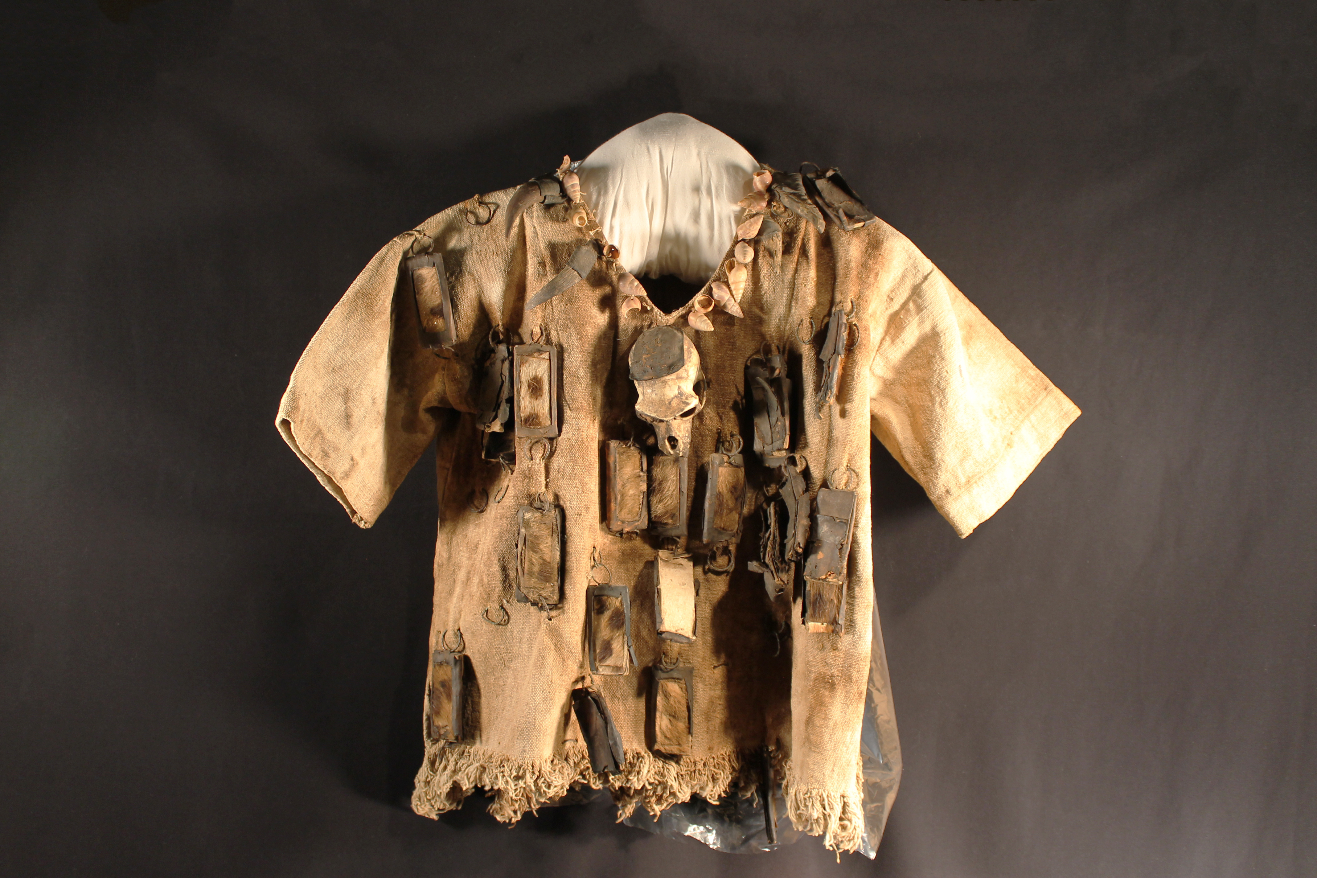 Cotton pullover short sleeved shirt with fringed bottom. Marine snail shells line the V-neck, a monkey skull in the middle, leather/hide pockets dangling off the shirt and shirt has been very well used consisting of sweat stains and dirt