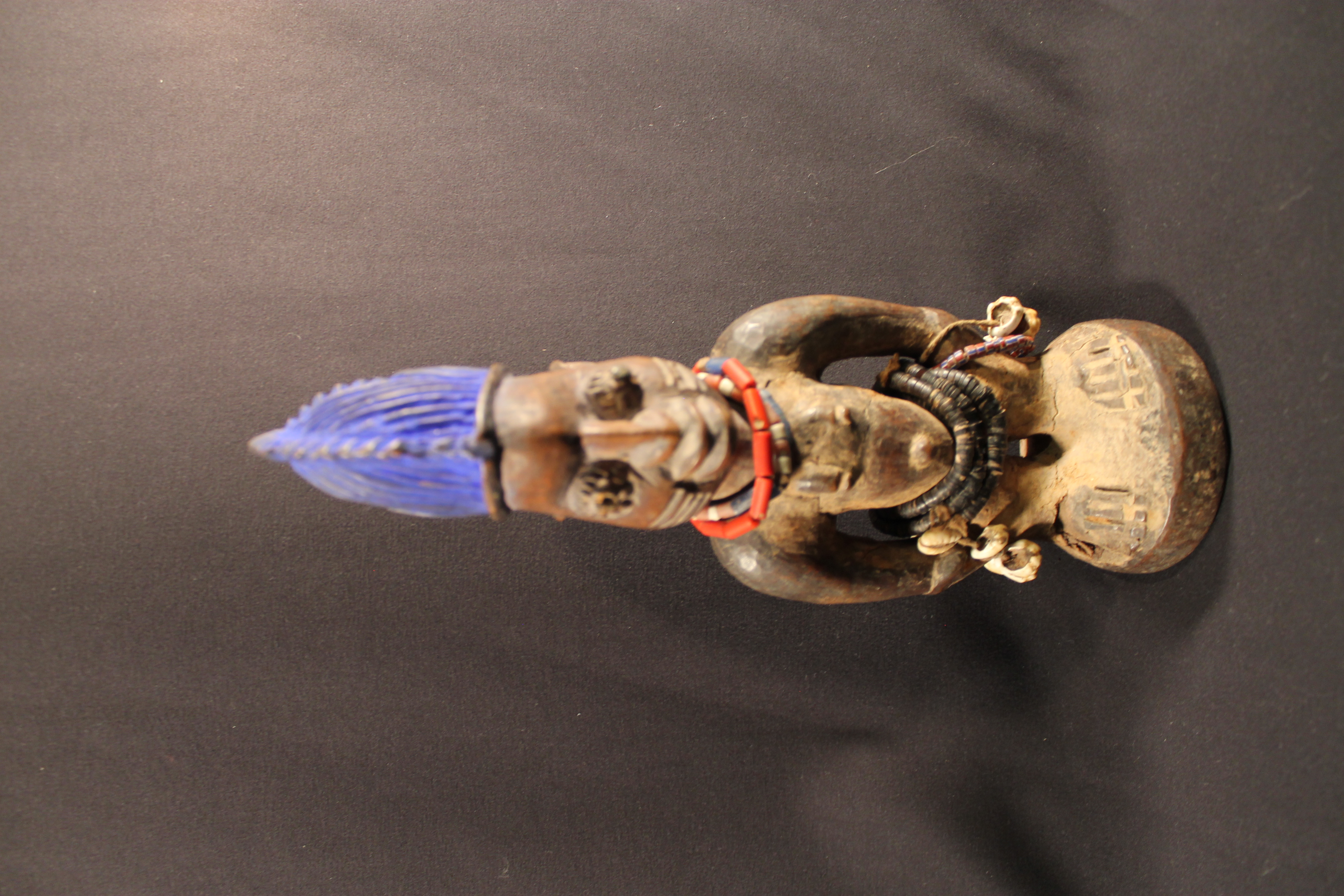 Figure is standing tall with a narrow face and bright blue hair. The figure is adorned with beaded necklaces and bracelets made of teeth. 