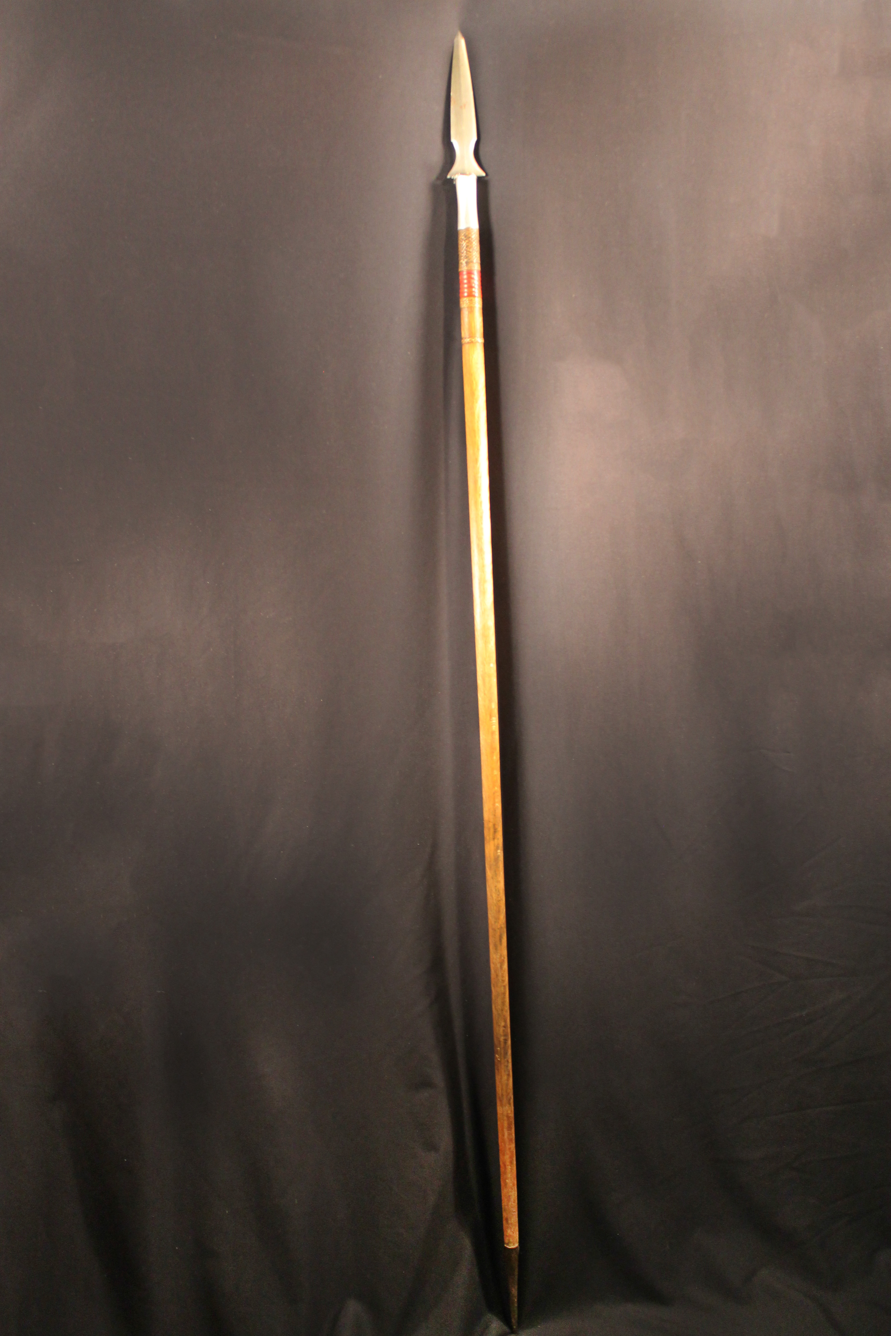 Spear made of light-colored wood with metal spear butt at bottom, below the spearhead is wrapped with red plants fibers and bands of braided plant material 