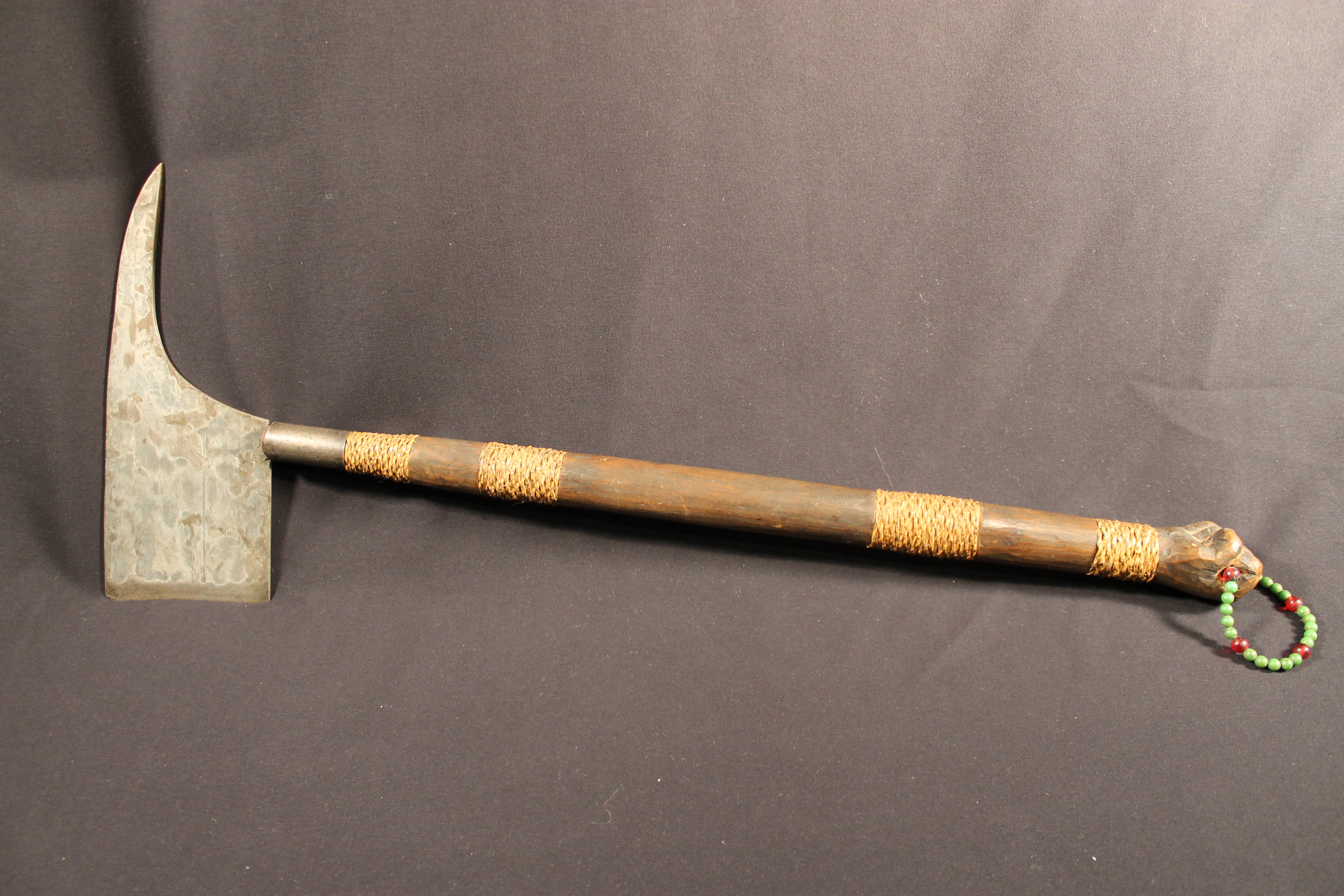 Metal axe head, front part is sharpened and back part is flat. Handle is a mixture of vegetal fibers and wood. Bottom of handle has a red and green beaded-like-bracelet looped through the end  