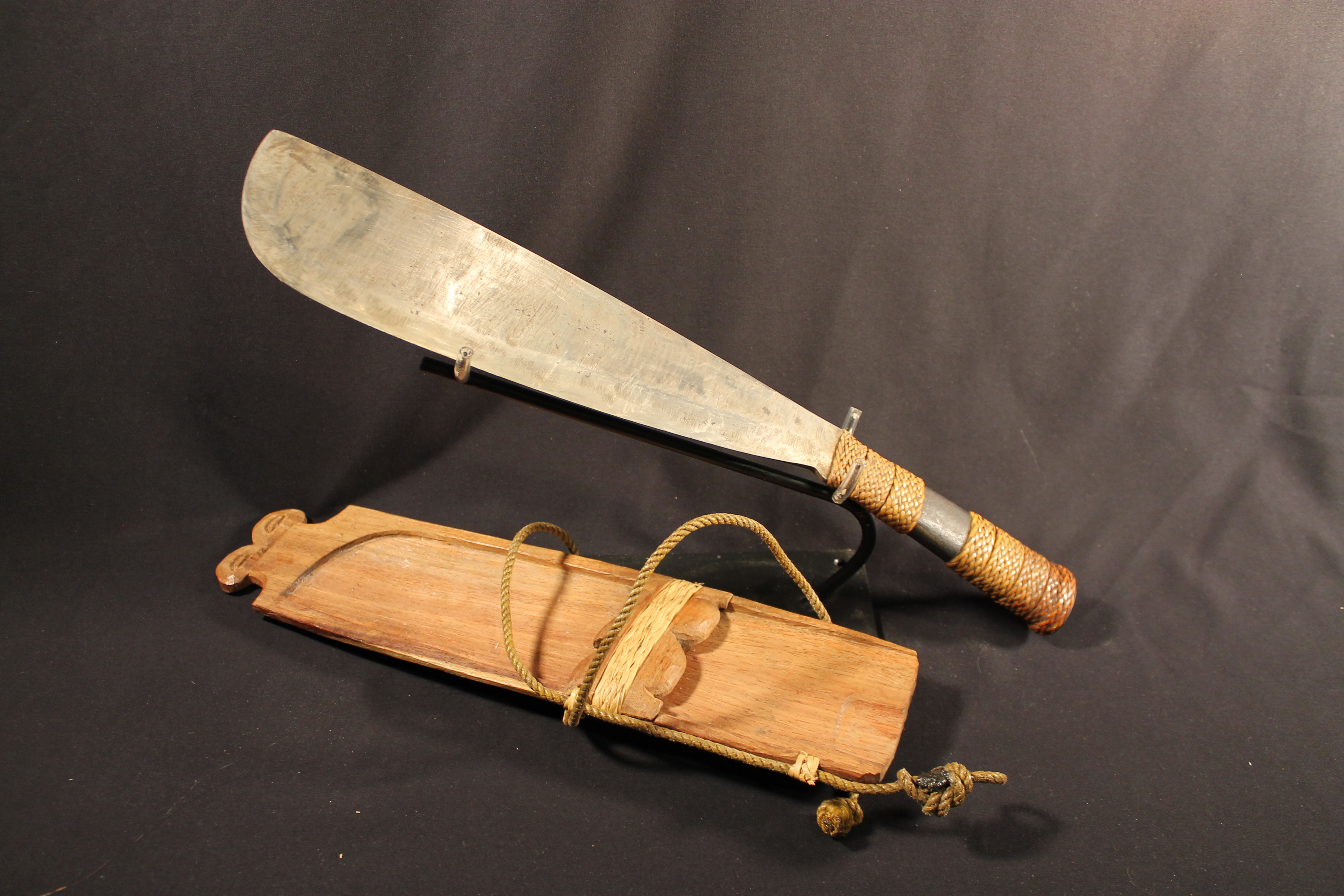 Knife with a wide blade and has several bands of braided plant fiber. Wooden knife sheath with carved decorations, band of braided plant fiber and an attached fiber rope 